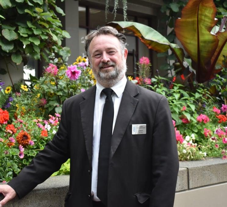 A man in a suit and tie stands proudly in front of a vibrant flower garden, showcasing a blend of elegance and nature