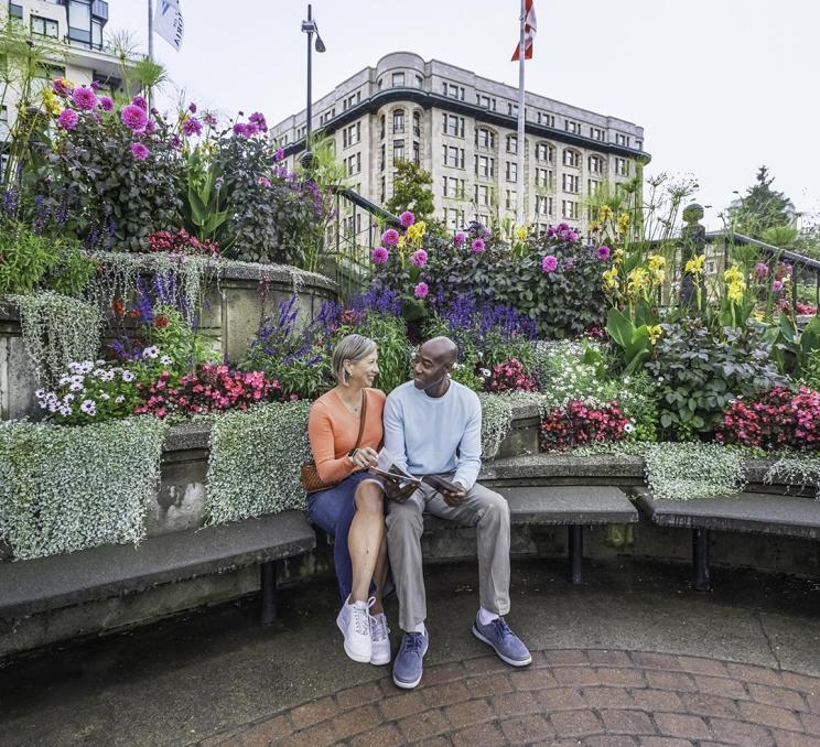 A couple reads a Victoria Vacation Guide while seated amidst some flowers outside of the Victoria Visitor Centre in Victoria, BC
